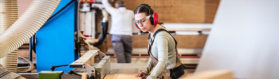 A student with safety equipment works on a saw table in a woodworking class